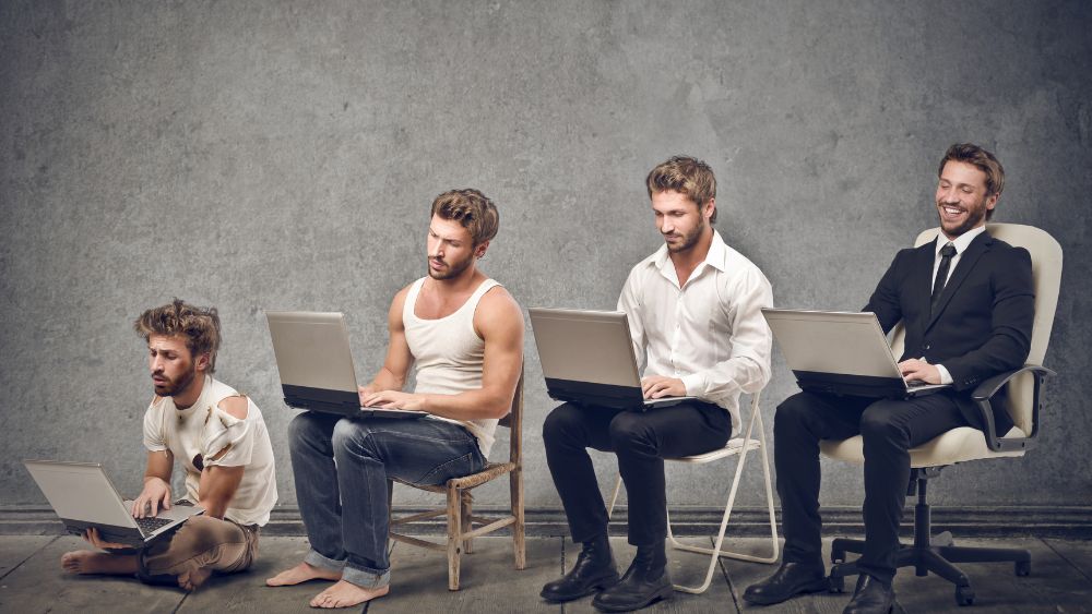 Four men at a laptop. From left to right each man looks progressively more comfortable and successful.